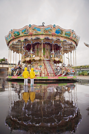 Two sweet children, boy brothers, watching carousel in the rain, wearing yellow raincoats, summertime, reflection on the wet groundの写真素材