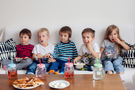Five sweet kids, friends, sitting in living room at home, watching TV and eating popcornの写真素材