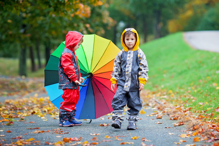 Two adorable children, boy brothers, playing in park with colorful rainbow umbrella on a rainy autumn dayの写真素材