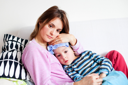 Young mother, holding her little sick boy, wet cloth on his forehead, trying to decrease his temperatureの写真素材