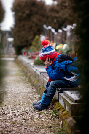 Sad little boy, sitting on a grave in a cemetery, feeling sad and cryingの写真素材