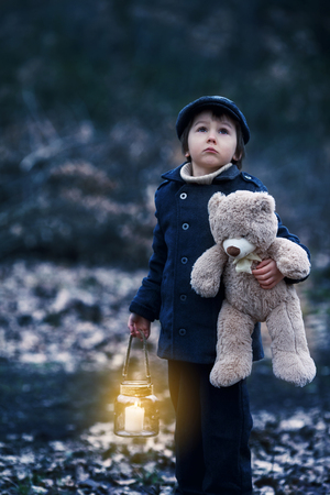 Cute little child, preschool boy, holding lantern and teddy bear, walking in a dark forestの写真素材