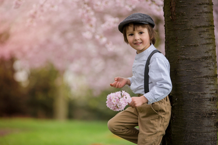 Beautiful young preschool boy, standing in a cherry blossom garden, holding flower, vintage outfit, smiling happily, spring childhood happiness conceptの写真素材