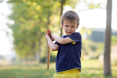 Angry little boy, holding sword, glaring with a mad face at the camera, outdoors in the parkの写真素材