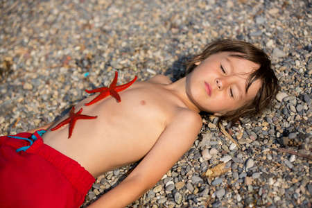 Adorable little boy, lying in the sand on the beach, two red starfishes lying down on his belly,...