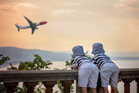 Two little children, boy brothers, looking at landing airplane in the sky over the sea on sunset and dreaming of becoming a pilotsの写真素材