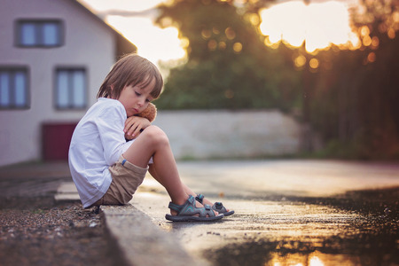 Sad little boy, sitting on the street in the rain, hugging his teddy bear, summertime on sunsetの写真素材