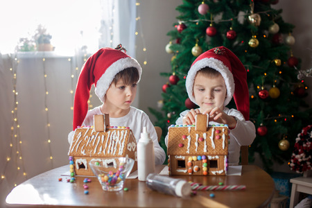 Two sweet boys, brothers, making gingerbread cookies house, decorating at home in front of the Christmas tree, child playing and enjoying, Christmas conceptの写真素材