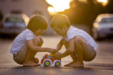 Two sweet children, boy brothers, playing with car toys on the street in village on sunset, summertimeの写真素材