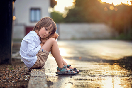 Sad little boy, sitting on the street in the rain, hugging his teddy bear, summertime on sunsetの写真素材