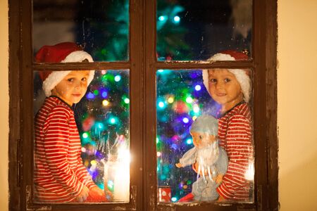 Two children, boys, sitting on a window at night, one holding toy, other playing on phone, waiting for Christmasの写真素材