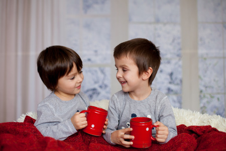 Cute children, boys, sitting in a big chair in pajamas, drinking tea and enjoying Christmas holidaysの写真素材