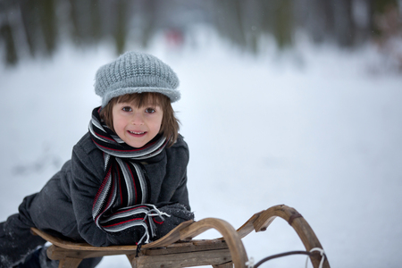 Cute little boy, lying down on sledge, smiling at camera, playing in the snow in winterの写真素材