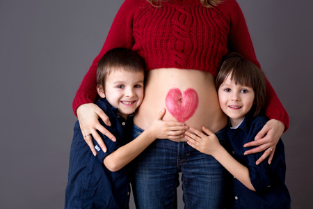 Beautiful preschool children, brothers, hugging their pregnant mom, happily expecting to be big brothers. Red heath drawn on mothers belly, child kissing her little bellyの写真素材