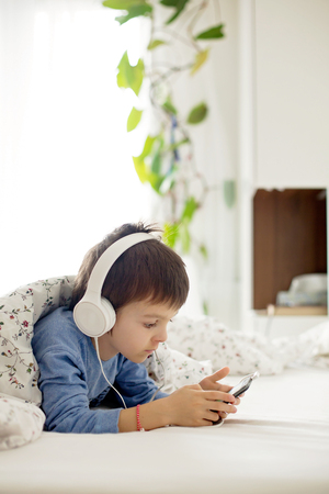 Little boy in pajama bed, playing on phone, listening music on headphonesの写真素材