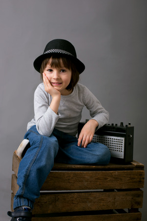 Cute boy with black hat, sitting on a wooden tray, holding old retro radio, smiling, isolated on gray backgroundの写真素材