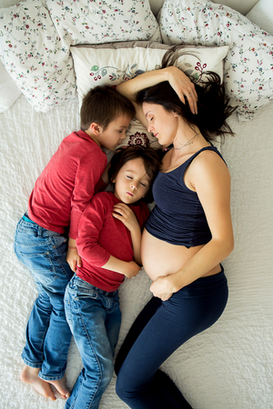 Beautiful pregnant young mother and her two children, boys, lyign in bed at homeの写真素材