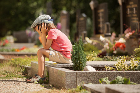 Sad little boy, sitting on a grave in a cemetery, feeling sad and cryingの写真素材