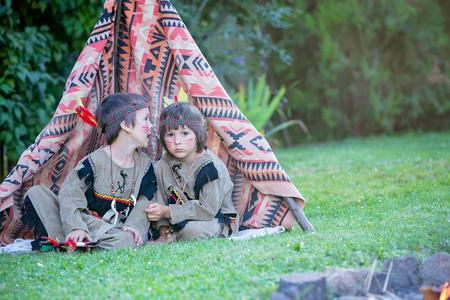Cute boy dressed in traditional  native american costume sitting by a tent outdoors in the parkの写真素材