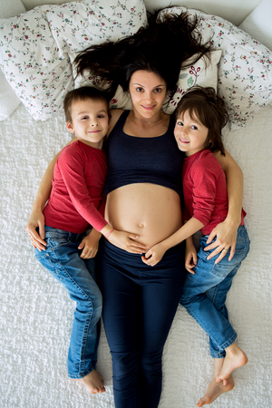 Beautiful pregnant young mother and her two children, boys, lying in bed at homeの写真素材