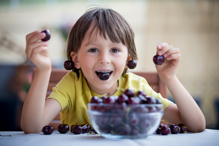 Cute little boy, eating sweet cherry outdoors, sitting on tableの写真素材