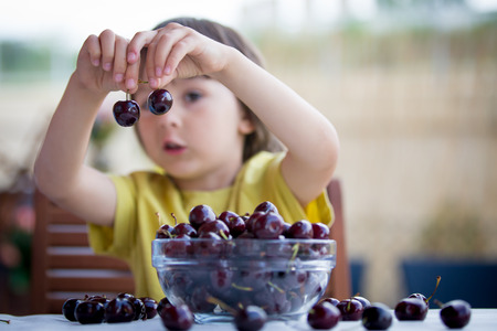 Cute little boy, eating sweet cherry outdoors, sitting on tableの写真素材