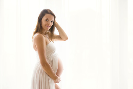 Portrait of young pregnant attractive woman, standing by the window, dressed in white dress, isolated imageの写真素材