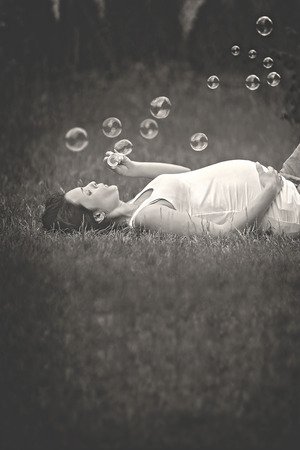 Beautiful young pregnant woman, lying down in the grass, making soap bubbles in gardenの写真素材