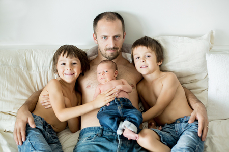 Father of three boys, preschool, toddler and newborn baby, sitting in bed, hugging togetherの写真素材
