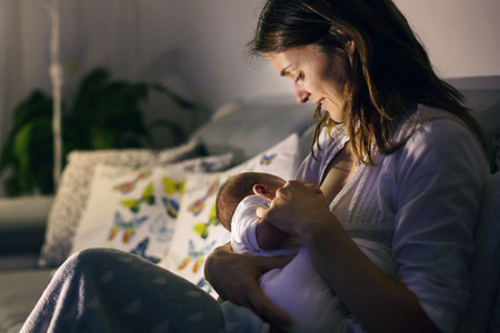 Young beautiful mother, breastfeeding her newborn baby boy at night, dim light. Mom breastfeeding...