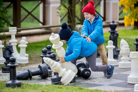 Two children, boy brothers, playing chess with huge figures in the park on the ground, autumn time. Childhood happiness concept, kids playing in the park fall timeの写真素材