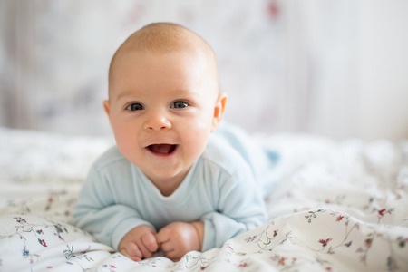 Adorable baby boy in white sunny bedroom in winter morning. Newborn child relaxing in bed. Family morning at home. Newborn kid during tummy time smiling happily at homeの写真素材