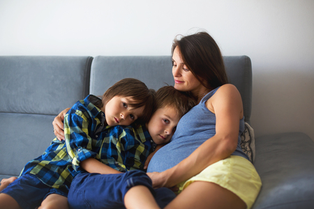 Portrait of beautiful pregnant mother, hugging with love and tenderness her precious children at homeの写真素材