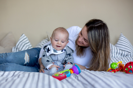 Cute little baby boy, playing with his mom at home in bed with lots of colorful toys for babies and...