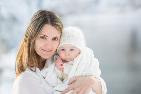 Beautiful mother in white dress and cute baby boy in knitted onesie, having taken their beautiful winter outdoor portrait on a sunny winter snowy dayの写真素材