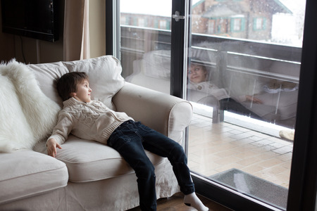 Preschool boy, sitting by the window in living room, looking at a snowy landscape outdoors, winter snowing weatherの写真素材