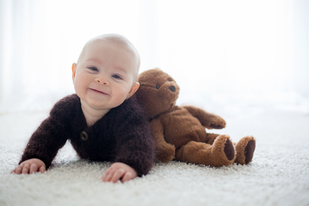 Little cute baby boy, dressed in handmade knitted brown teddy bear overall, playing at home in sunny bedroomの写真素材