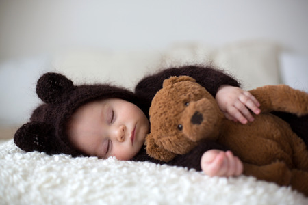 Sweet little baby boy, dressed in handmade knitted brown soft teddy bear overall, sleeping cozy at home in sunny bedroom with lots of teddy bears around himの写真素材