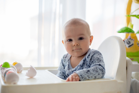 Cute little toddler boy, playing with plastic eggs, sitting in a white chair in a sunny living room, white around him, baby boy smiling happilyの写真素材
