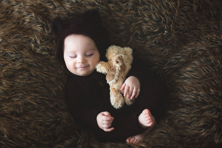 Little baby boy with knitted brown jumpsuit as a bear, sleeping at home on a furry blanket, smiling with happinessの写真素材