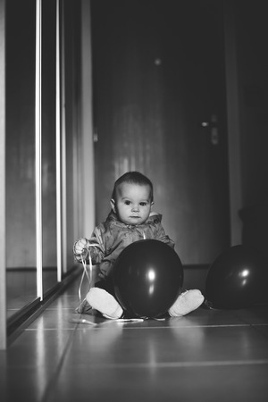 Adorable baby boy, toddler kid, sitting in the entrance at home on the floor, holding red balloonsの写真素材