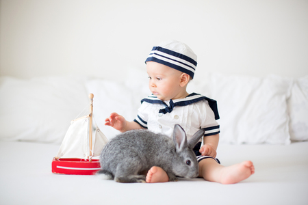 Cute baby boy, dressed in marine clothes, playing with wooden boat and little cute bunny in bedroomの写真素材