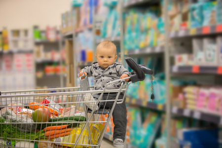 Toddler baby boy, sitting in a shopping cart in grocery store, smiling and eating bread  while mommy is shoppingの写真素材