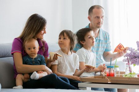 Beautiful young family with three children, eating pizza at home and watching TV on a sunday