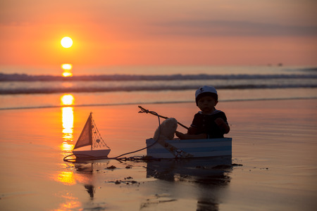 Cute baby child, sweet boy, playing with boat, teddy bear and fishes on sunset at the edge of the ocean, beautiful sunset, reflectionの写真素材
