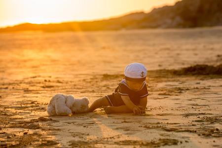 Cute baby child, sweet boy, playing with boat, teddy bear and fishes on sunset at the edge of the ocean, beautiful sunset, reflectionの写真素材