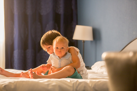 Two sibling children, baby boy and his older brother, sitting on bed in sunny bedroom, playing together, back lightの写真素材