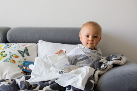 Sick little toddler boy, sitting on a couch in living room with a box of tissuesの写真素材