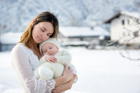 Beautiful mother in white dress and cute baby boy in knitted onesie, having taken their beautiful winter outdoor portrait on a sunny winter snowy dayの写真素材