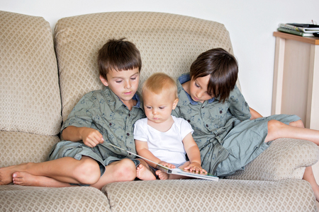 Three boys, siblings, sitting on a sofa and reading a book in living room, summertime
